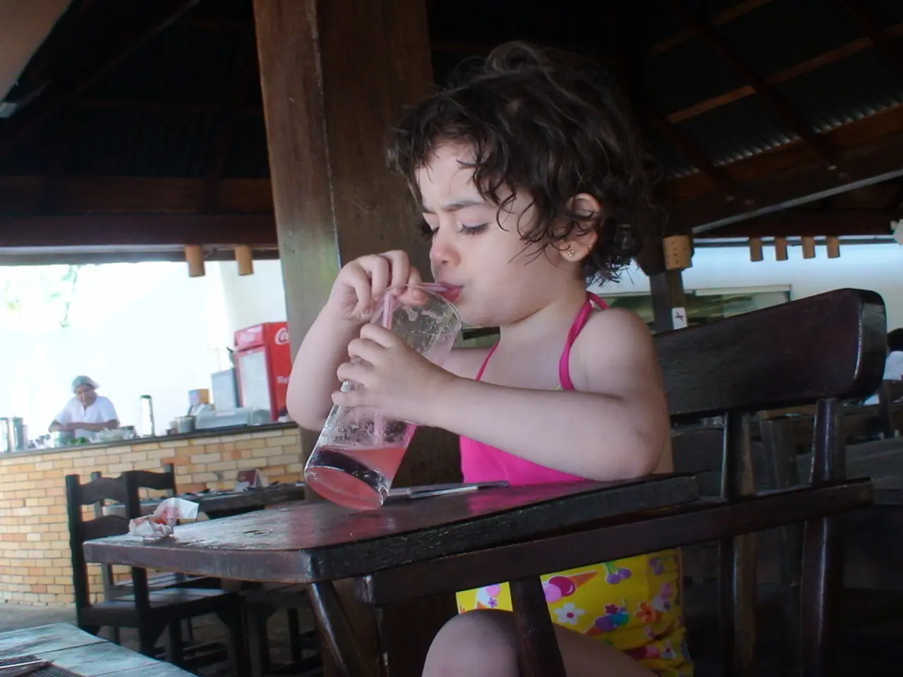 She is sitting on a chair. She is drinking a juice. There is a table. There is a tissue,pen on a...